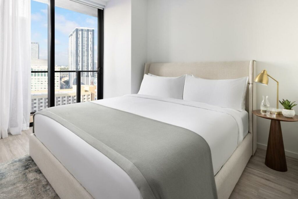 Quarto do The Elser Hotel Miami – An All-Suite Hotel com cama de casal do lado direito da imagem com uma mesa com luminária do lado esquerdo da cama. Representa hotéis para Brasileiros em Miami.