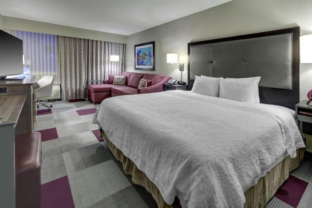 Quarto do Hampton Inn Miami-Coconut Grove/Coral Gables com cama de casal do lado direito da imagem em cada lado da cama uma cômoda com luminária em frente a cama uma cômoda com TV em cima do lado direito da cama um pequeno sofá e em frente uma mesa com cadeira. Representa hotéis para Brasileiros em Miami.