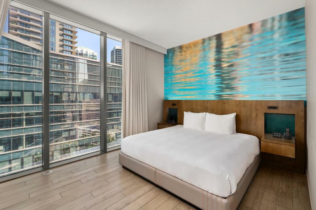 Imagem do quarto do EastMiami com cama no centro do quarto em cada lado da cama uma cômoda e do lado esquerdo da cama uma janela panorâmica. Representa hotéis para Brasileiros em Miami.