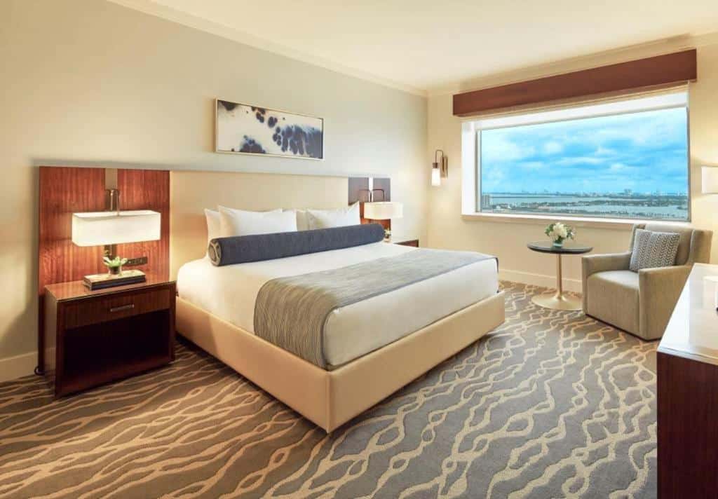 Quarto do InterContinental com cama de casal ao centro de carro, duas cômodas de madeira com luminária, com poltrona do lado esquerdo. Representa hotéis para Brasileiros em Miami.