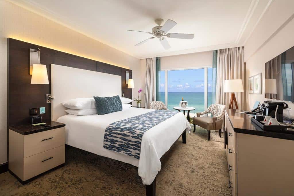 Imagem do quarto do The Palms Hotel & Spa com cama de casal do lado esquerdo da imagem com uma cômoda em cada lado da cama, em frente a cama uma cômoda com TV presa na parede, do lado esquerdo duas poltronas com mesa redonda atrás das poltrona uma janela panoramica com vista para a praia. Representa hotéis para Brasileiros em Miami.