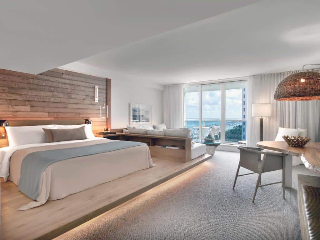 Imagem do quarto do 1 Hotel South Beach com cama de casal do lado esquerdo da imagem do lado esquerdo da cama uma sala de estar com um sofá e em frente uma mesa de madeira com cadeira. Representa hotéis para Brasileiros em Miami.
