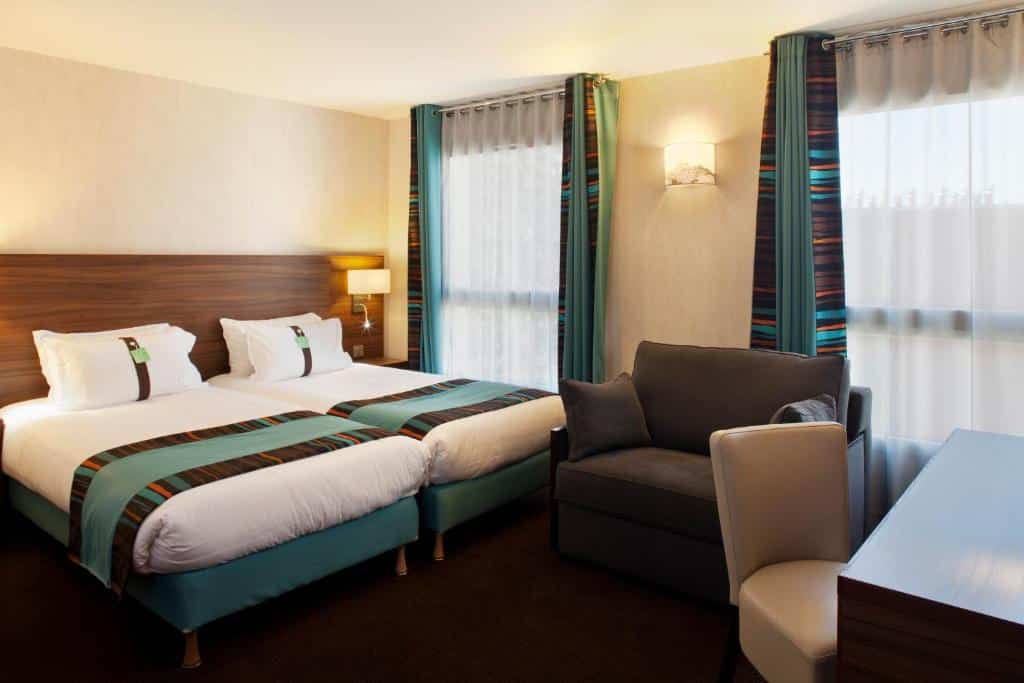 Quarto do Holiday Inn Paris Montmartre, um dos hotéis para família em Paris, com duas camas de solteiro, um sofá, uma escrivaninha com uma cadeira e janelas grandes com cortinas transparentes e listradas.