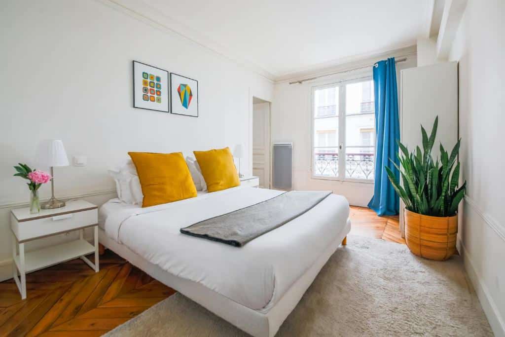 Quarto bem iluminado do Residence Bergère, um dos hotéis para família em Paris, com uma cama de casal, travesseiros amarelos, mesas de cabeceira, uma planta grande e uma janela com cortinas azuis.