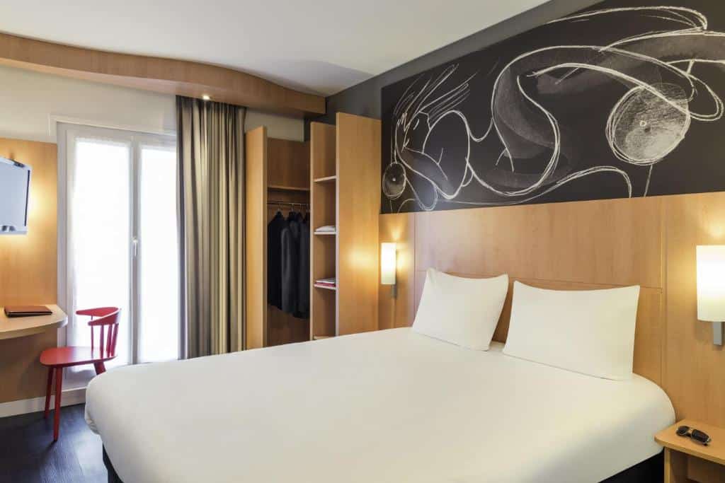 Quarto do ibis Paris Avenue d'Italie 13ème, um dos hotéis para família em Paris, com uma cama de casal, armário aberto, arte na parede, cadeira vermelha, escrivaninha, luminárias de parede e janela com cortinas.