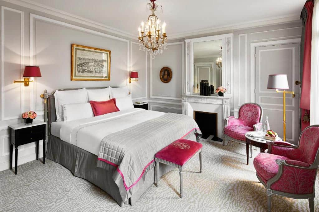 Quarto do Hôtel Plaza Athénée, um dos hotéis para família em Paris, sendo ele elegante com uma cama grande, poltronas, lustre e uma lareira, decorado em tons de branco, cinza e rosa.