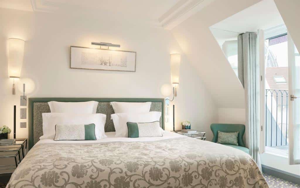 A imagem mostra um quarto do Le Burgundy Paris, com cama bem arrumada com roupas de cama estampadas em um quarto bem iluminado, duas mesinhas de cabeceira, uma poltrona e uma porta para uma pequena varanda.