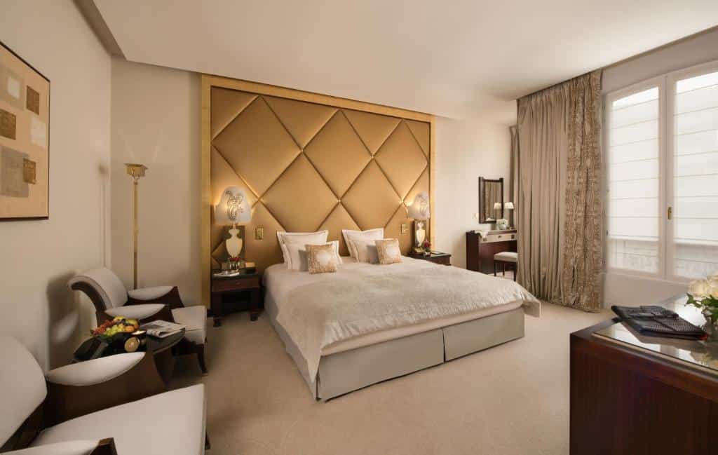 Quarto do Hôtel Barrière Le Fouquet's, com uma cama de casal, paredes bege, cabeceira dourada, duas cadeiras, uma escrivaninha, luminárias e uma grande janela com cortinas.