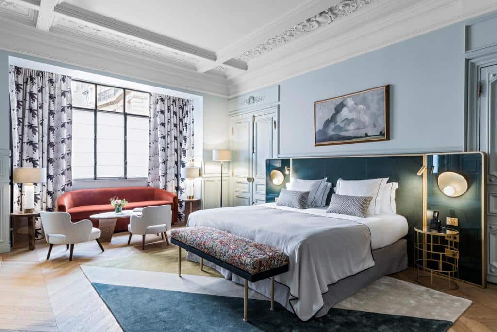 Quarto do Grand Powers Hotel, um dos hotéis para família em Paris, moderno com uma cama grande, um sofá vermelho, duas cadeiras, um banco e uma janela com cortinas estampadas.