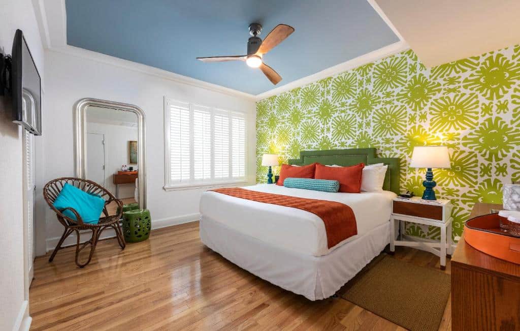 Quarto do Circa 39 Hotel Miami Beach com cama de casal no centro da imagem com uma cômoda em cada lado da cama com luminária, no teto no centro um ventilado, em frente a cama uma TV presa na parede e do lado esquerdo da cama uma cômoda e do lado direito uma cadeira com um espelho. Representa melhores hotéis em Miami.