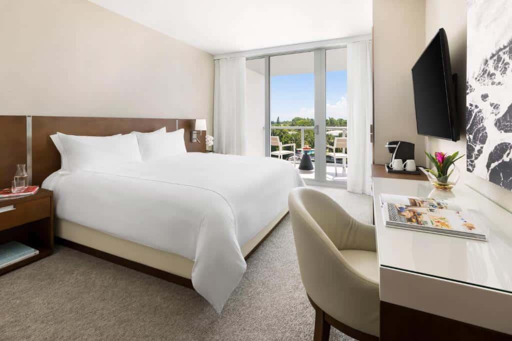 Quarto do The Altair Bay Harbor Hotel  com cama de casal do lado esquerdo da imagem, em cada lado da cama uma cômoda, em frente a cama uma cômoda com cadeira e uma TV presa na parede. Representa melhores hotéis em Miami.