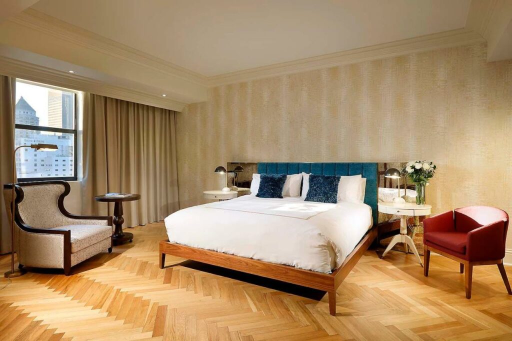 Imagem do quarto do Eurostars Langford com cama de casal no centro do quarto em cada lado da cama uma cômoda com luminária do lado direto uma poltrona e do lado esquerdo uma poltrona com uma mesa redonda de madeira ao lado. Representa melhores hotéis em Miami.