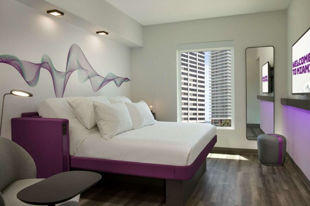 Quarto do Yotel Miami com cama de casal do lado esquerdo da imagem no centro do quarto em frente a cama uma TV presa na cidade e do lado direito da cama uma poltrona. Representa melhores hotéis em Miami.