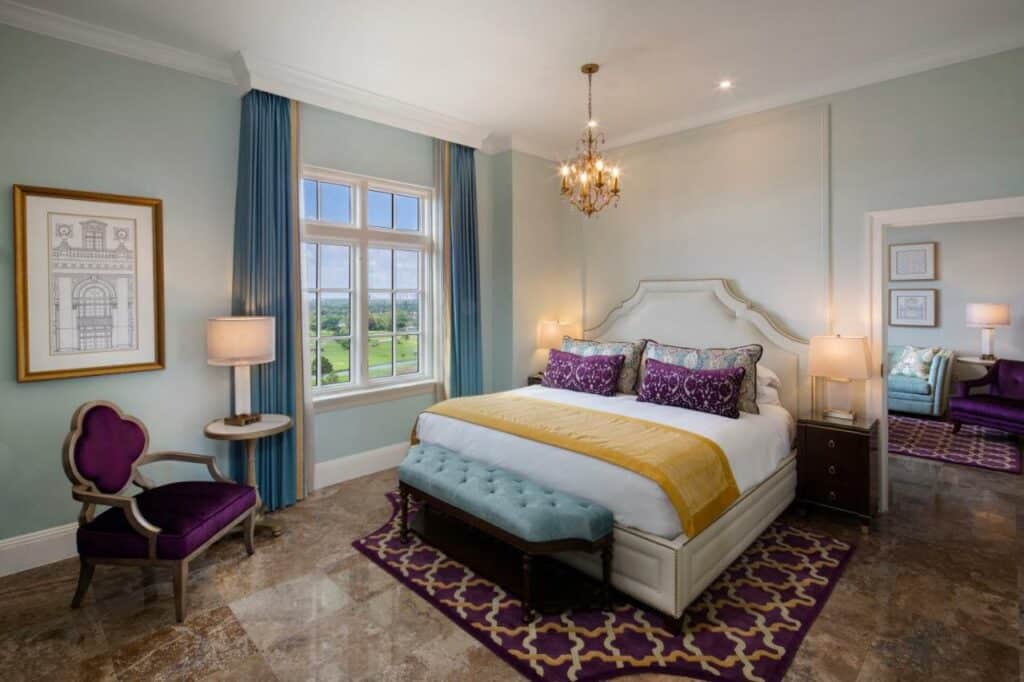 Quarto do Biltmore Hotel Miami Coral Gables com cama de casal do lado direito no centro do quarto com uma cômoda em cada lado com luminária, no pé da cama uma banco estofado e do lado esquerdo ao pé da cama uma poltrona e ao lado esquerdo da poltrona uma mesinha com luminária. Representa melhores hotéis em Miami.