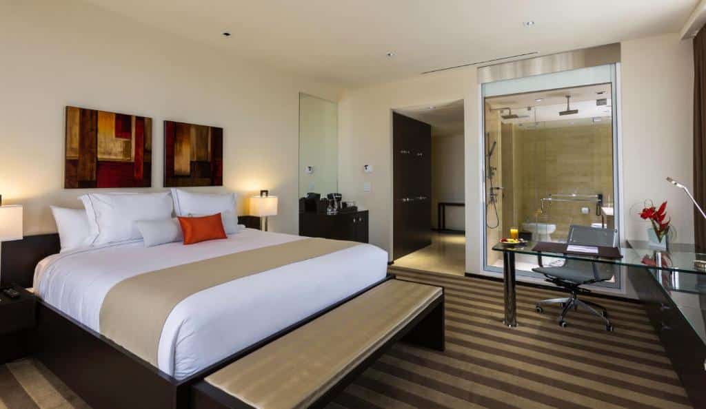 Quarto do EB Hotel Miami Airport com cama de casal do lado esquerdo da imagem com uma cômoda e luminária em cada lado da cama, no pé da cama um banco estofado do lado esquerdo a frente uma mesa com cadeiras. Representa melhores hotéis em Miami.
