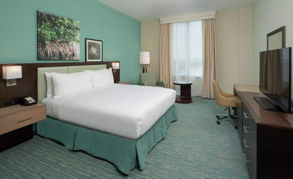 Quarto do DoubleTree by Hilton Miami Doral com cama de casal do lado esquerdo da imagem com uma cômoda em cada lado da cama, em frente a cama uma cômoda com TV em cima do lado esquerdo da cômoda uma mesa com cadeira e ao fundo uma mesa redonda com poltrona. Representa melhores hotéis em Miami.