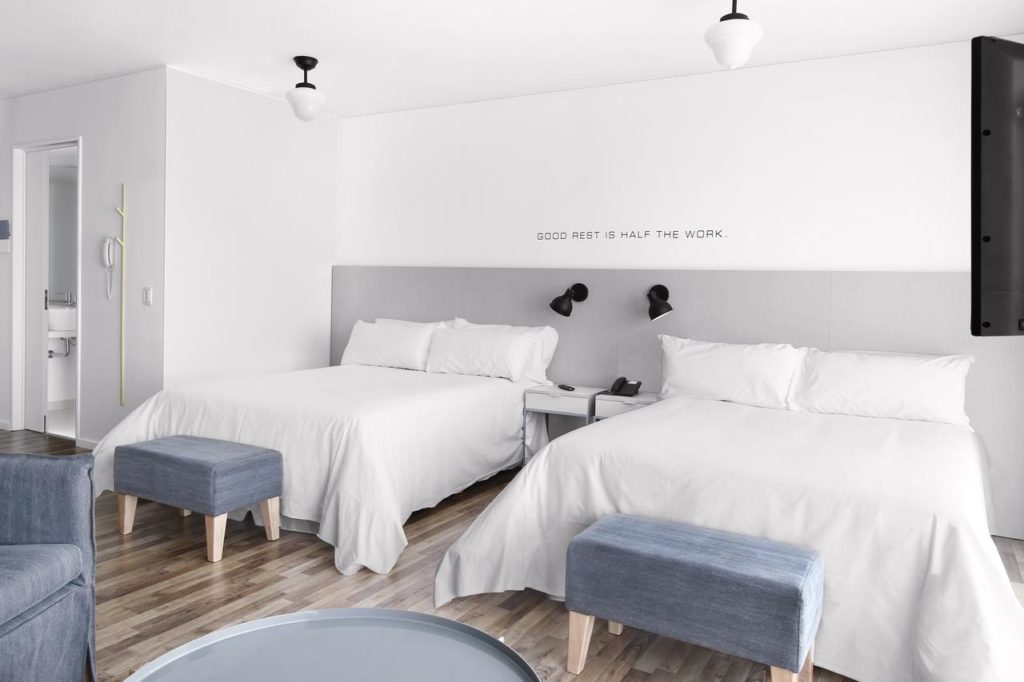 Quarto minimalista do CH Madero Urbano Suites em Porto Madero, com duas camas de casal, cabeceiras cinza, roupa de cama branca e uma citação na parede acima das camas, para representar onde ficar em Buenos Aires.