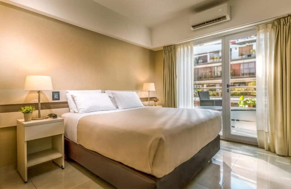 Uma cama bem arrumada em um quarto no Up Recoleta Hotel, bem iluminado com mesas de cabeceira, luminárias e uma porta de vidro que dá para uma varanda, para representar onde ficar em Buenos Aires.