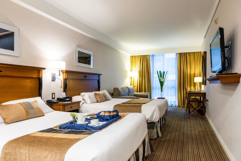 Quarto no Blank Hotel Recoleta com duas camas de solteiro, uma bandeja com café da manhã, um sofá, TV na parede e janela grande com cortinas amarelas, para representar onde ficar em Buenos Aires.