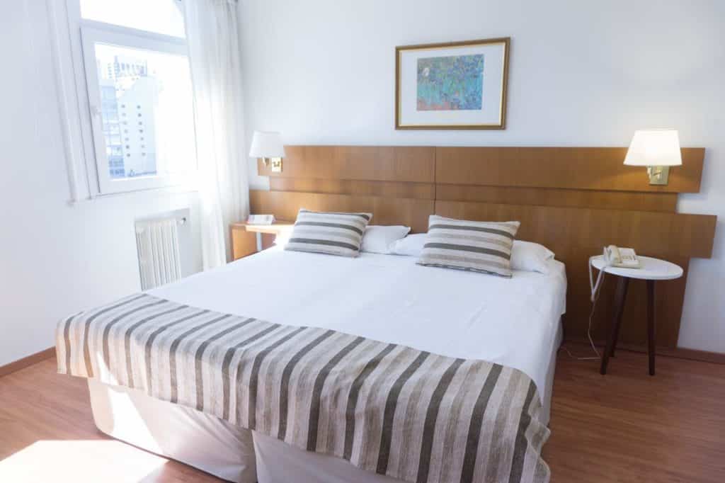Uma cama de casal bem arrumada com roupa de cama listrada em um quarto bem iluminado, duas mesas de cabeceira, luminárias e uma janela, no Hotel Bristol, para representar onde ficar em Buenos Aires.