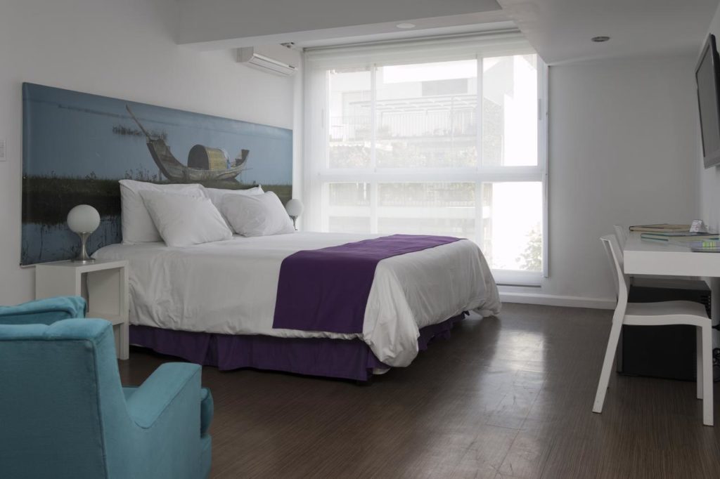 Um quarto moderno no Infinito Hotel, com uma cama de casal, detalhes em roxo, uma cadeira azul, uma escrivaninha e uma grande janela com cortinas transparentes, para representar onde ficar em Buenos Aires.