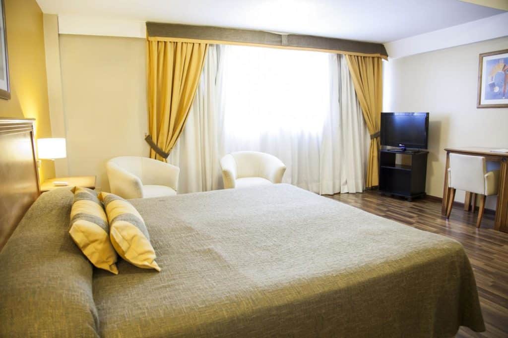 Um quarto no Tritone Hotel com uma cama grande, duas cadeiras, uma TV, uma pequena escrivaninha e cortinas amarelas cobrindo uma janela, para representar onde ficar em Beunos Aires.