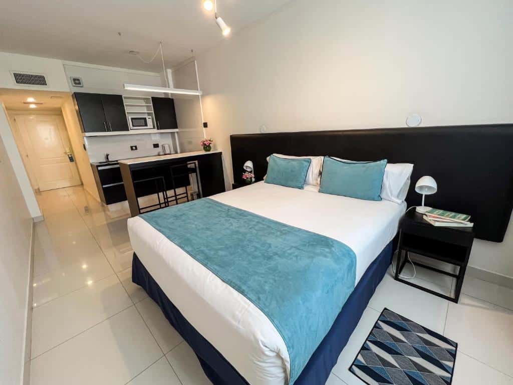 Um estúdio moderno com uma cama, cabeceira preta, roupa de cama azul-petróleo, área de cozinha e piso de azulejos brancos, no Cyan Recoleta Hotel, para representar onde ficar em Buenos Aires.