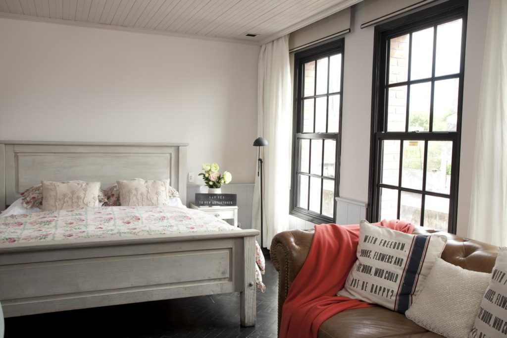 Um quarto com uma cama de madeira, roupa de cama floral, um sofá de couro marrom e janelas grandes com cortinas brancas, na Casa Chic Palermo Soho, para representar onde ficar em Buenos Aires.