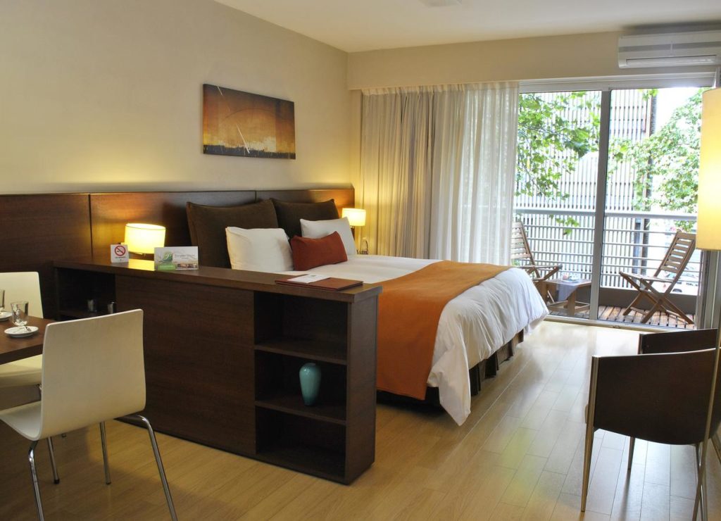 Quarto moderno no Palermo Suítes Buenos Aires, com uma cama de casal, escrivaninha e varanda com duas cadeiras e uma pequena mesa, com luz do dia do lado de fora.