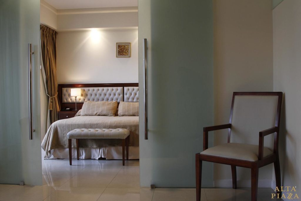 Um quarto no Alta Piazza - Casa di Appartamenti, com cama, banco, poltrona, mesa de cabeceira e arte na parede, visto através de portas de vidro fosco, para representar onde ficar em Buenos Aires.