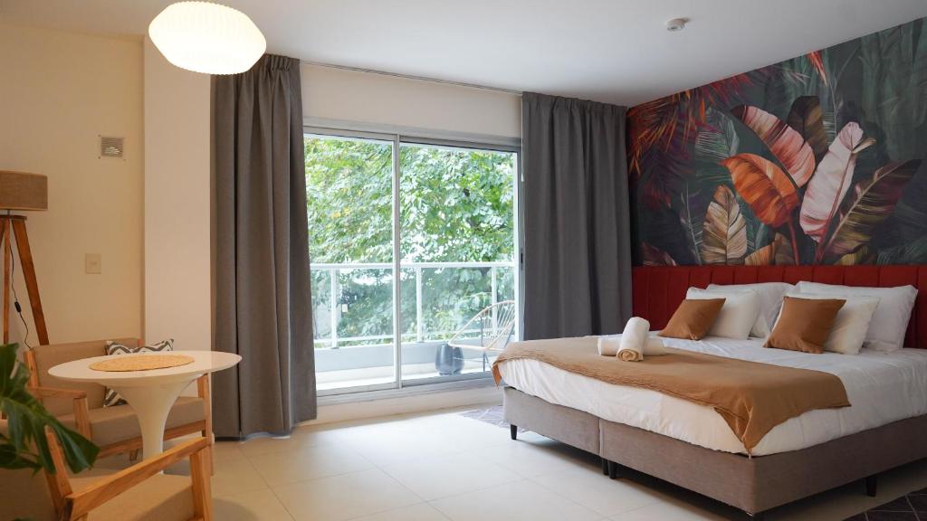 Um quarto moderno com uma cama grande, parede de destaque com tema tropical, mesa redonda, cadeira e portas de vidro deslizantes para uma varanda, na CASARICA Aparts, para representar onde ficar em Buenos Aires.