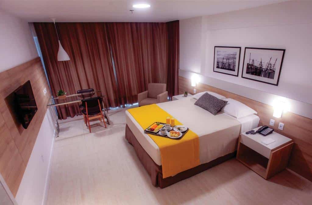 Quarto no Gran Mareiro Hotel na Praia do Futuro, com uma cama de casal, escrivaninha, cadeira, TV e bandeja de café da manhã sobre a cama, com cortinas marrons fechadas, para representar onde ficar em Fortaleza.
