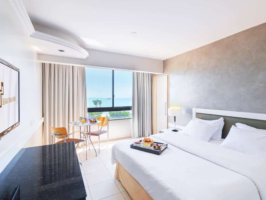Quarto no Seara Praia Hotel na Praia do Meireles, com uma cama grande, mesa e cadeiras ao lado de uma janela, vista para o mar e uma bandeja de café da manhã sobre a cama, para representar onde ficar em Fortaleza.