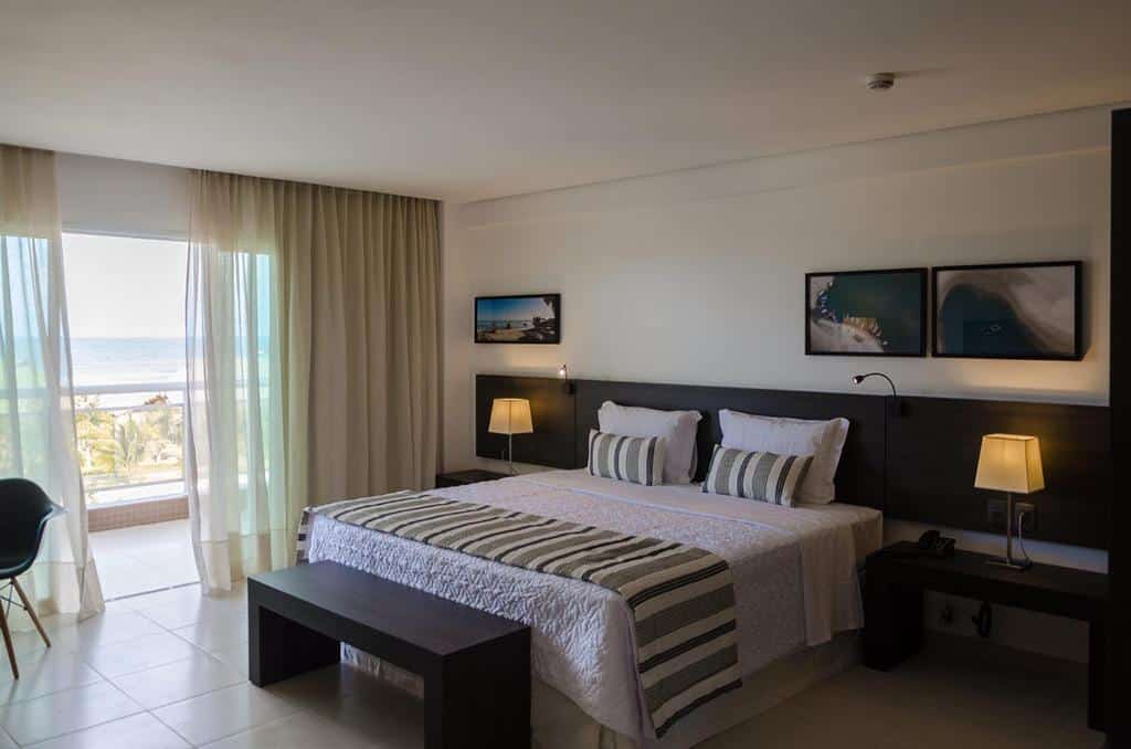 Um quarto no Crocobeach Hotel na Praia do Futuro, com uma cama grande, duas luminárias, um banco, dois quadros na parede e uma varanda com vista para o mar, para representar onde ficar em Fortaleza.