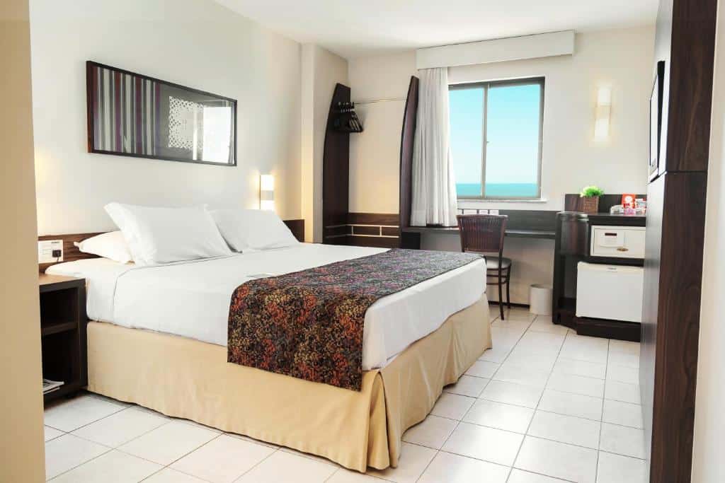 Um quarto no Hotel Sonata de Iracema, na Praia de Iracema, com uma cama de casal, escrivaninha, cadeira, frigobar, espelho de parede e uma janela com vista para o mar, para representar onde ficar em Fortaleza.