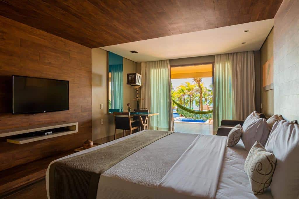 Quarto moderno no Carmel Charme Resort em Aquiraz, com uma cama king-size, escrivaninha, rede no pátio e vista para a piscina e palmeiras do lado de fora, para representar onde ficar em Fortaleza.