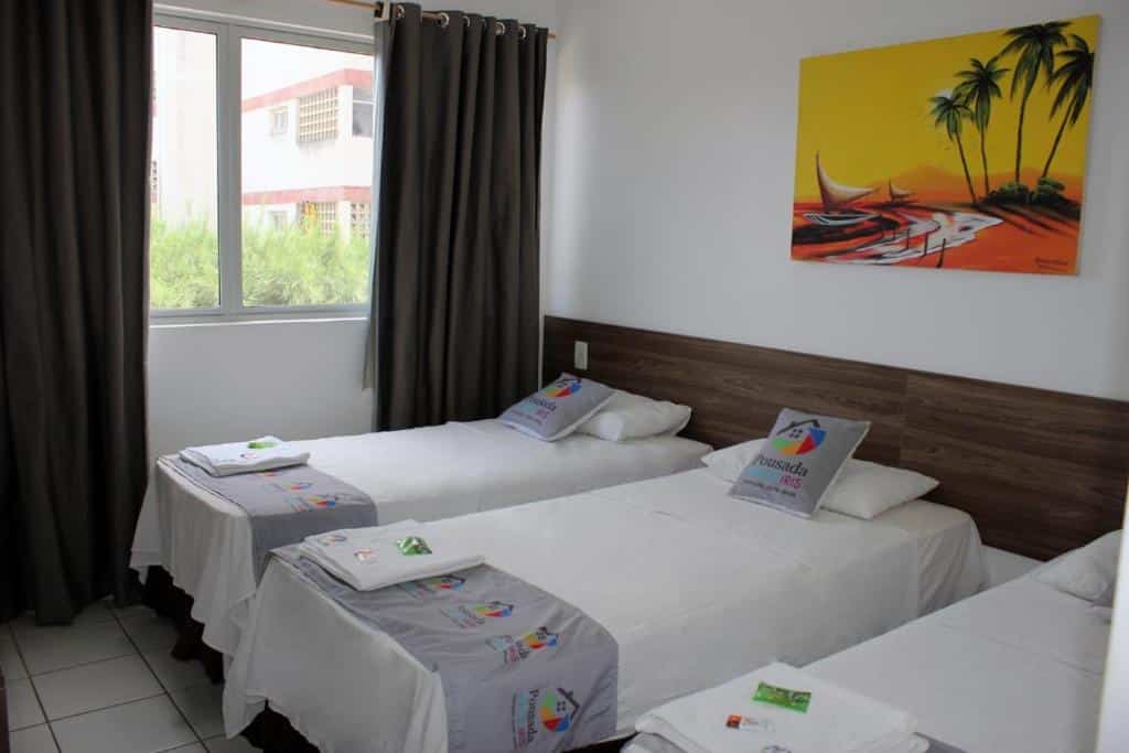 Um quarto na Pousada Arco Iris na Praia do Futuro, com três camas de solteiro, roupa de cama branca, toalhas dobradas, uma janela e uma pintura tropical na parede, para representar onde ficar em Fortaleza.
