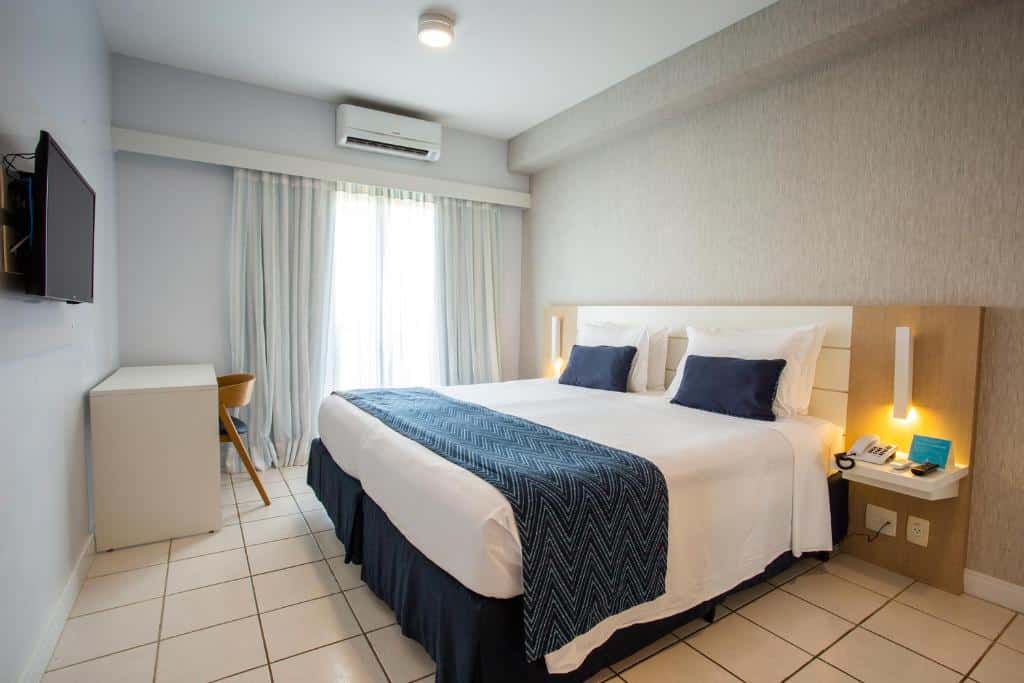 Quarto no Beach Park Resort em Aquiraz, com uma cama de casal, detalhes em azul, TV na parede, escrivaninha e luminárias de cabeceira, com luz natural da janela, para representar onde ficar em Fortaleza.
