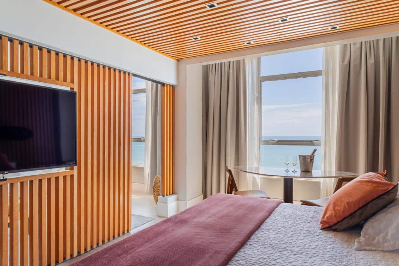 Um quarto moderno do Praiano Hotel com uma cama perto da janela, TV em uma parede de ripas de madeira, duas cadeiras perto de uma mesa pequena e vista para o mar.