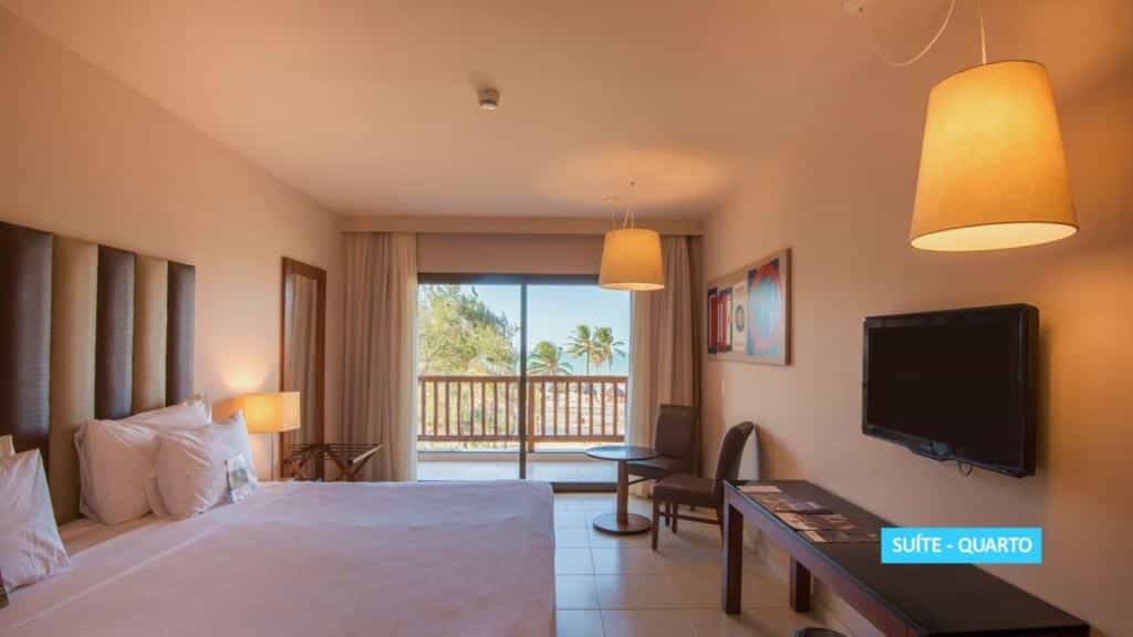 Quarto na Vila Galé Resort em Cumbuco com uma cama grande, escrivaninha, cadeiras, TV na parede e varanda com vista para as palmeiras, para representar onde ficar em Fortaleza.