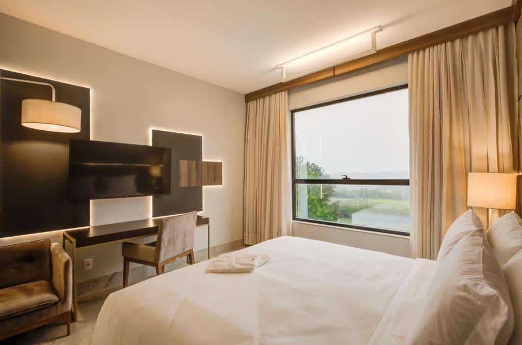 Foto do quarto do DoubleTree by Hilton, ilustrando o post sobre Onde ficar em Foz do Iguaçu. Há uma cama box de casal na direita, e de frente há um painel com TV e uma mesa escrivaninha, cadeira e poltrona. No fundo há uma janela com cortinas beges.