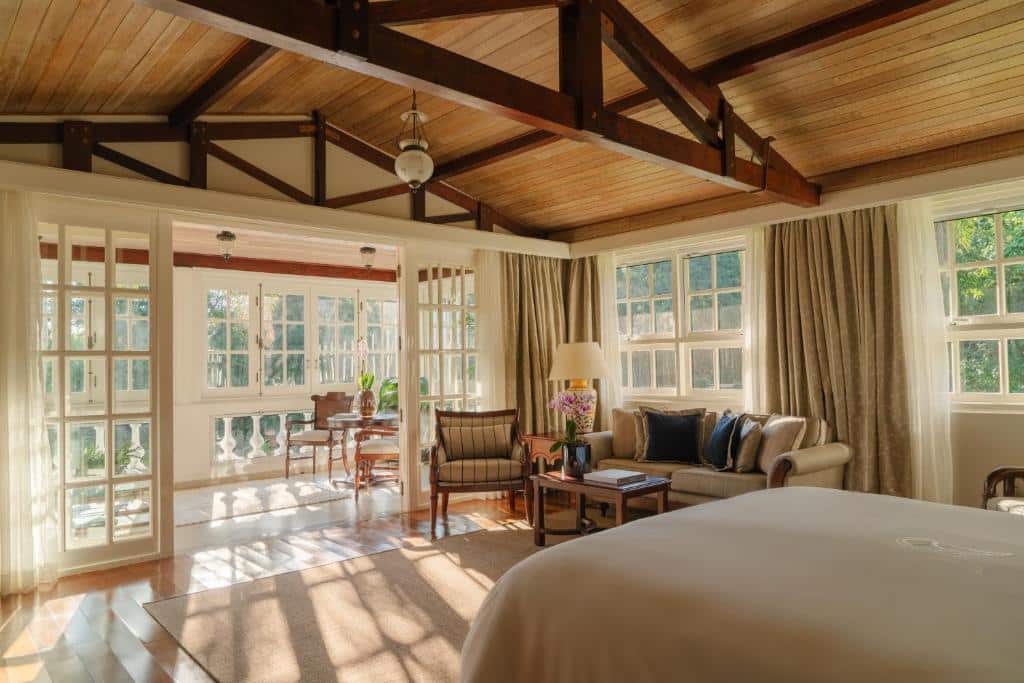 Foto do quarto do Belmond Hotel das Cataratas, ilustrando o post sobre Onde ficar em Foz do Iguaçu. O espaço é grande, há uma cama box de casal no canto inferior direito. Na frente da cama há um sofá com poltrona e mesa de centro. Já mais ao fundo há uma porta de vidro com outro espaço de descanso que dá acesso para a varanda da suíte.