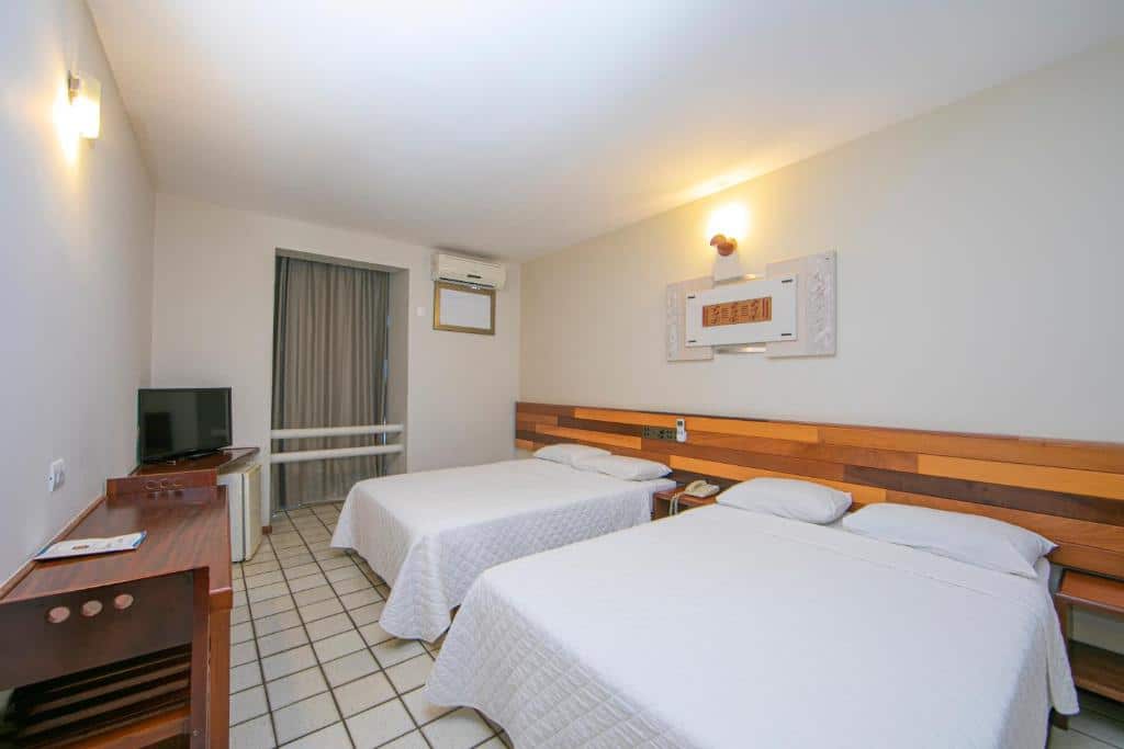 Foto do quarto do Falls Galli Hotel, ilustrando o post sobre Onde ficar em Foz do Iguaçu. Há duas camas de casal encostadas na parede á direita, e na frente delas há uma mesa com TV, frigobar e cadeira. Há uma janela no quarto e ar-condicionado.