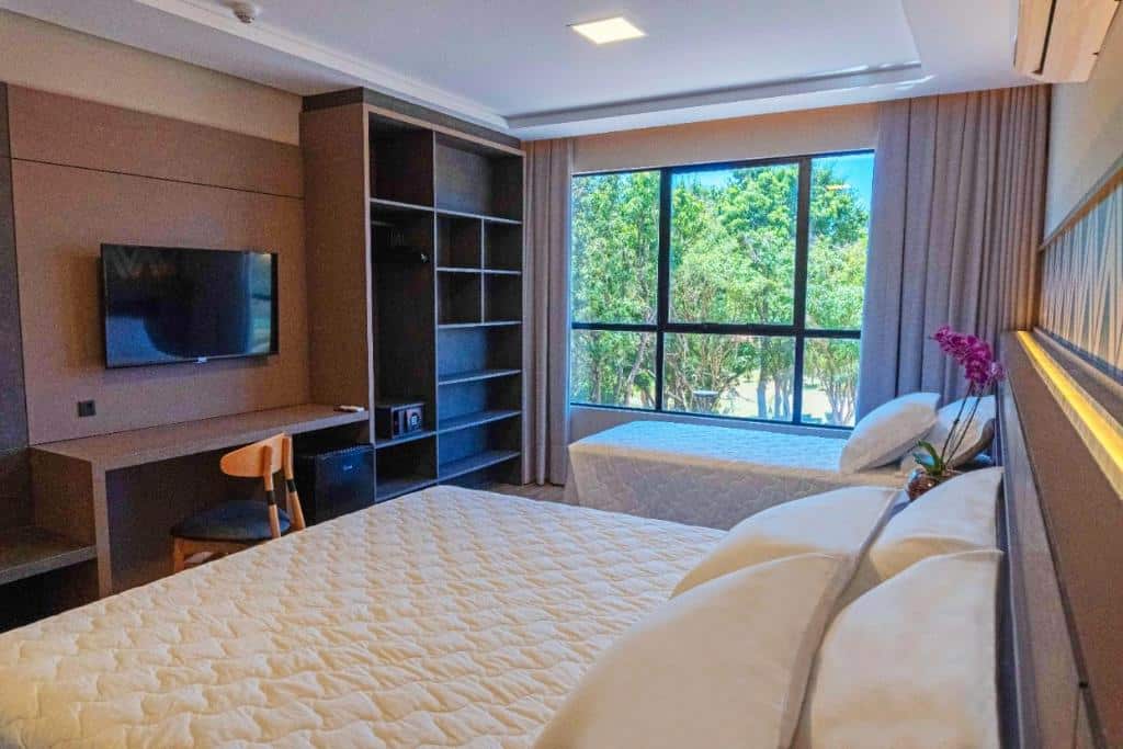 Foto do quarto do Complexo Eco Cataratas Resort, ilustrando o post sobre Onde ficar em Foz do Iguaçu. Há uma cama box de casal em primeiro plano, e outra de solteiro ao seu lado, no fundo da foto. E ao lado da cam de solteiro há uma janela. Na esquerda há uma escrivaninha, TV e estante com prateleiras vazias.
