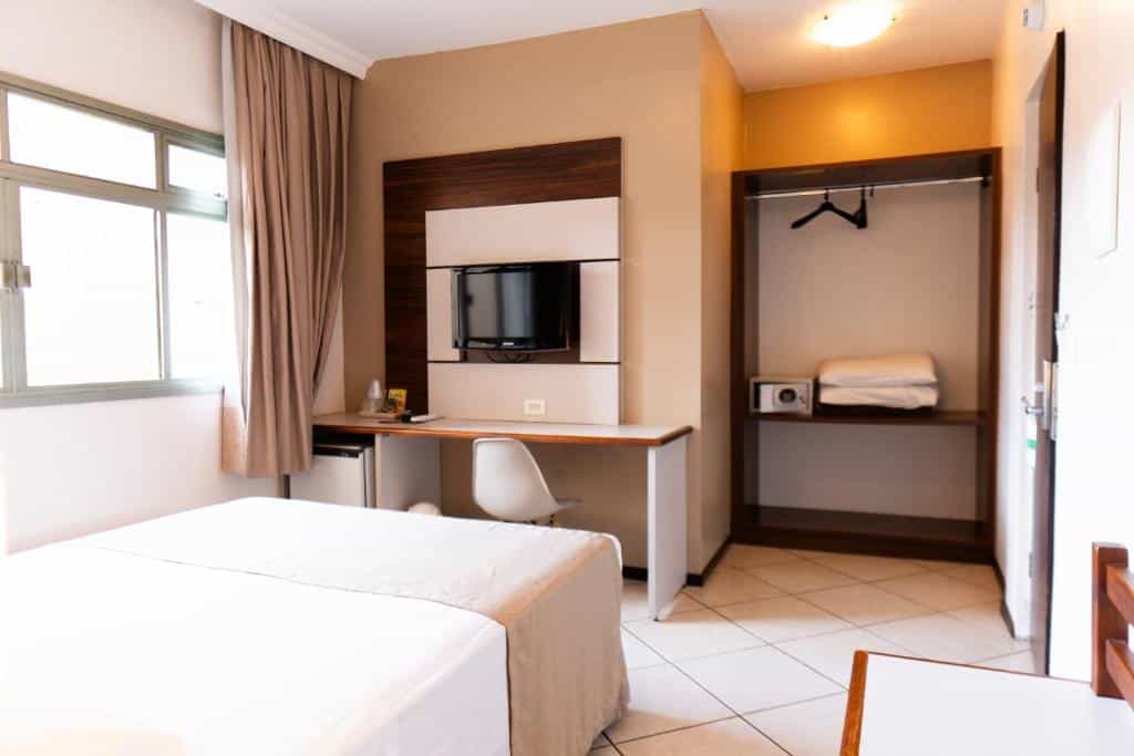 Foto do quarto do San Rafael Comfort Class Hotel, ilustrando o post sobre Onde ficar em Foz do Iguaçu. Vemos o quarto na visão de quem está sentado na cama. A cama está no canto esquerdo, e na frente há uma escrivaninha, frigobar e uma TV. Na direita há uma estante com cofre e travesseiros. E mais para a direita há uma porta.