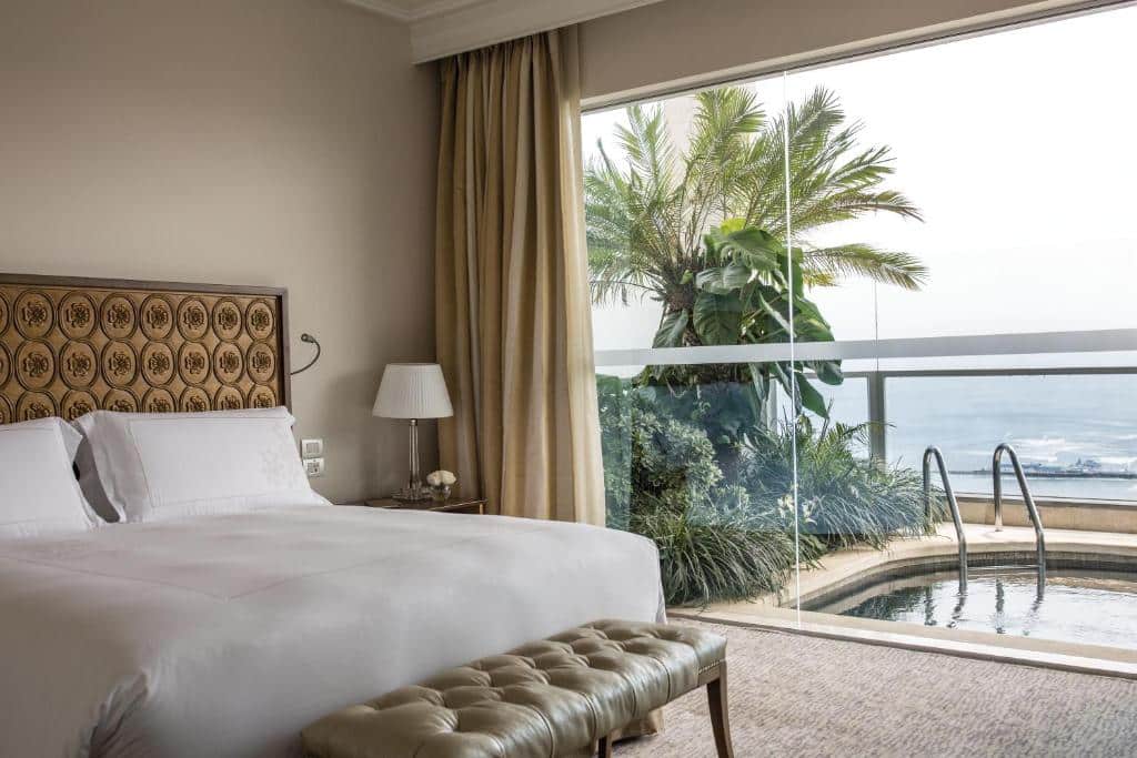 Quarto de hotel com uma cama de casal, banco e uma janela com vista para uma varanda com uma pequena piscina e vista para o mar. Um dos hotéis onde ficar em Lima.