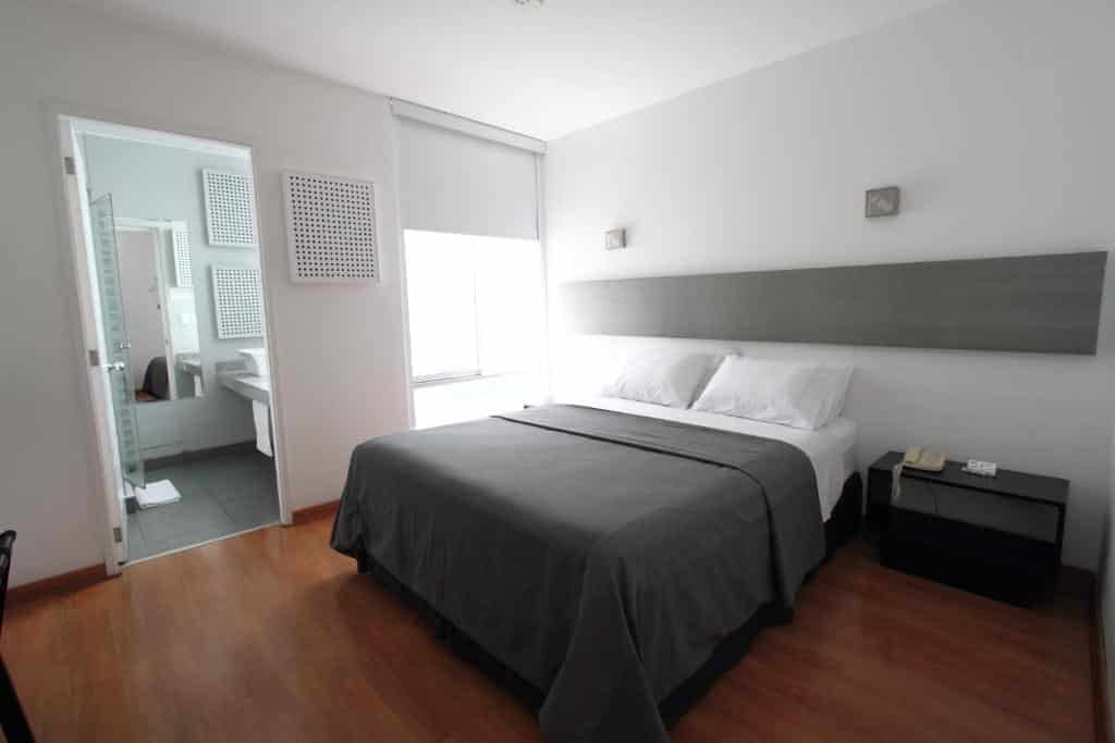 Quarto de um dos lugares onde ficar em Lima, o 3B Barranco's. Um quarto moderno com uma cama de casal, roupa de cama cinza, mesa de cabeceira, piso de madeira e um banheiro anexo.