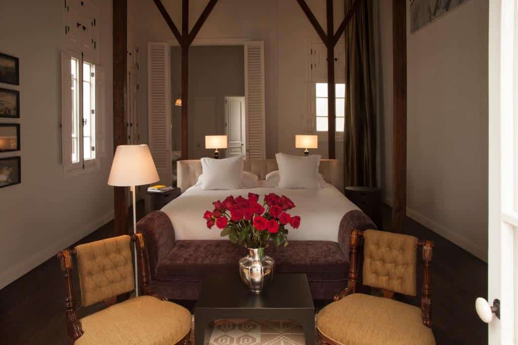 Duas camas de solteiro com roupa de cama branca, luminárias de cabeceira, um vaso de rosas vermelhas e duas cadeiras em um quarto aconchegante e bem iluminado.