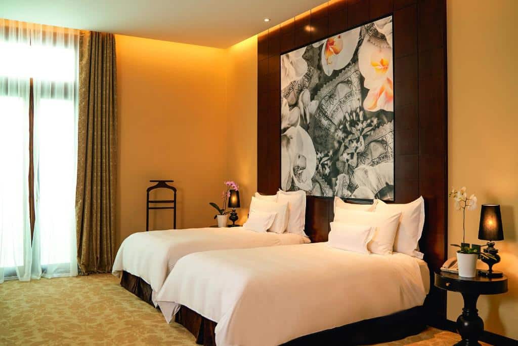 Quarto de hotel com duas camas de casal, roupa de cama branca, luminárias de cabeceira, arte floral na parede e janela grande com cortinas.