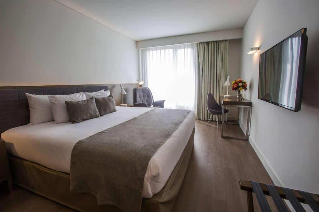 Quarto de hotel moderno com uma cama grande, escrivaninha, cadeira, poltrona, TV na parede e janela com cortinas.